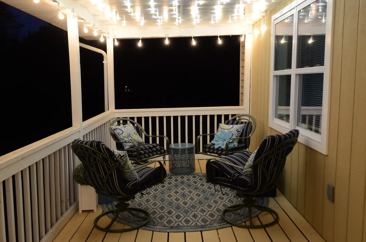 An inviting outdoor lounge setup with string lights overhead, comfortable seating, decorative pillows, and a textured rug. The cozy arrangement is ideal for relaxing evenings, stargazing, or socializing in an intimate, resort-like atmosphere.