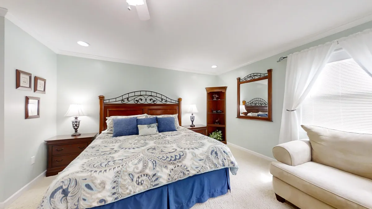 A serene king suite featuring a king-sized bed with decorative bedding, nightstands with lamps on each side, a large window dressed with sheer curtains letting in gentle natural light, and a wooden bookshelf. The space exudes calm and invites relaxation, perfect for unwinding after a busy day.