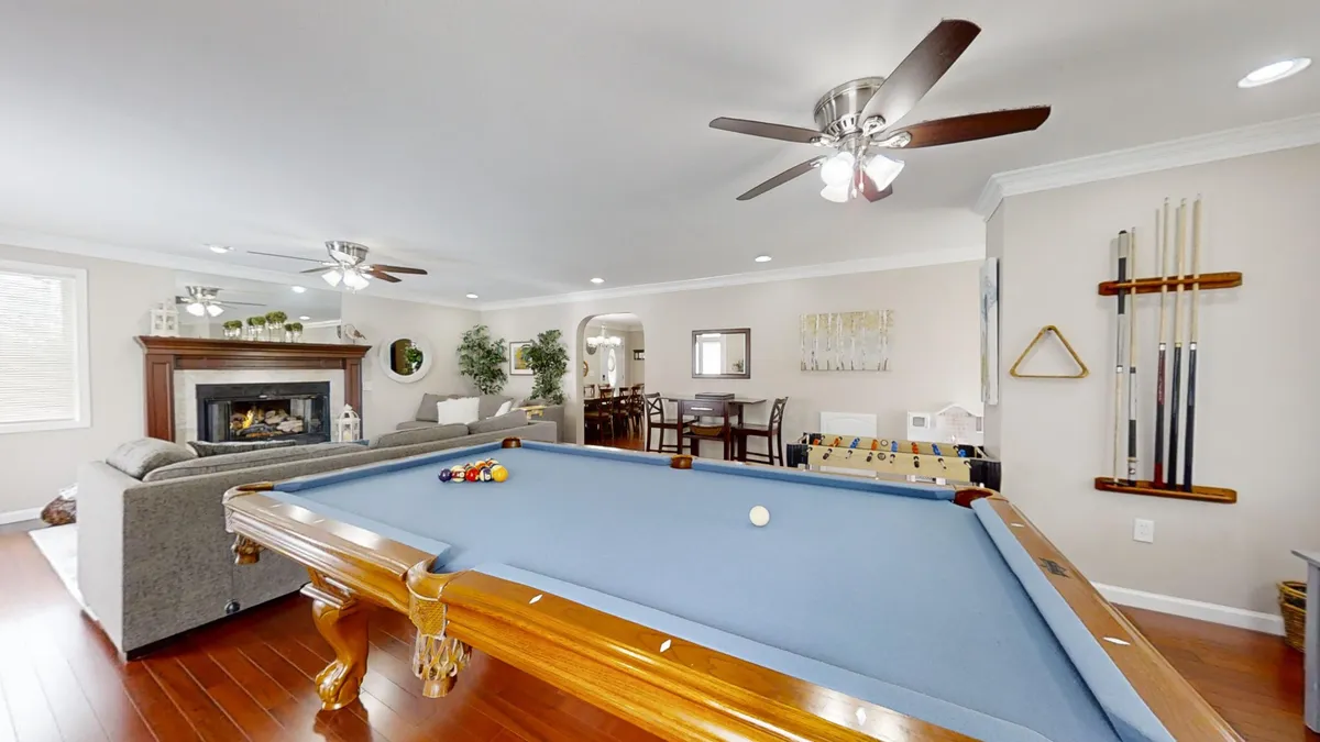 A spacious, open living area featuring a pool table in the foreground, a cozy couch nearby, and a warm fireplace along one wall. Ceiling fans and large windows add to the inviting, casual atmosphere, making it ideal for both game play and comfortable relaxation.