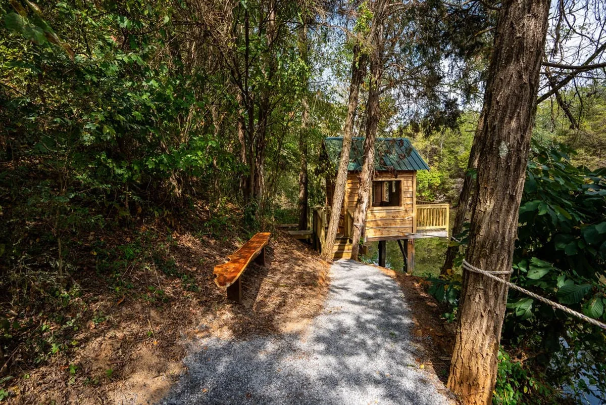 A charming treehouse nestled among tall trees, with a winding pathway leading up to it. There is a wooden bench positioned to take in the peaceful natural surroundings, and the entire setting is enveloped in greenery, evoking a sense of adventure and serenity.