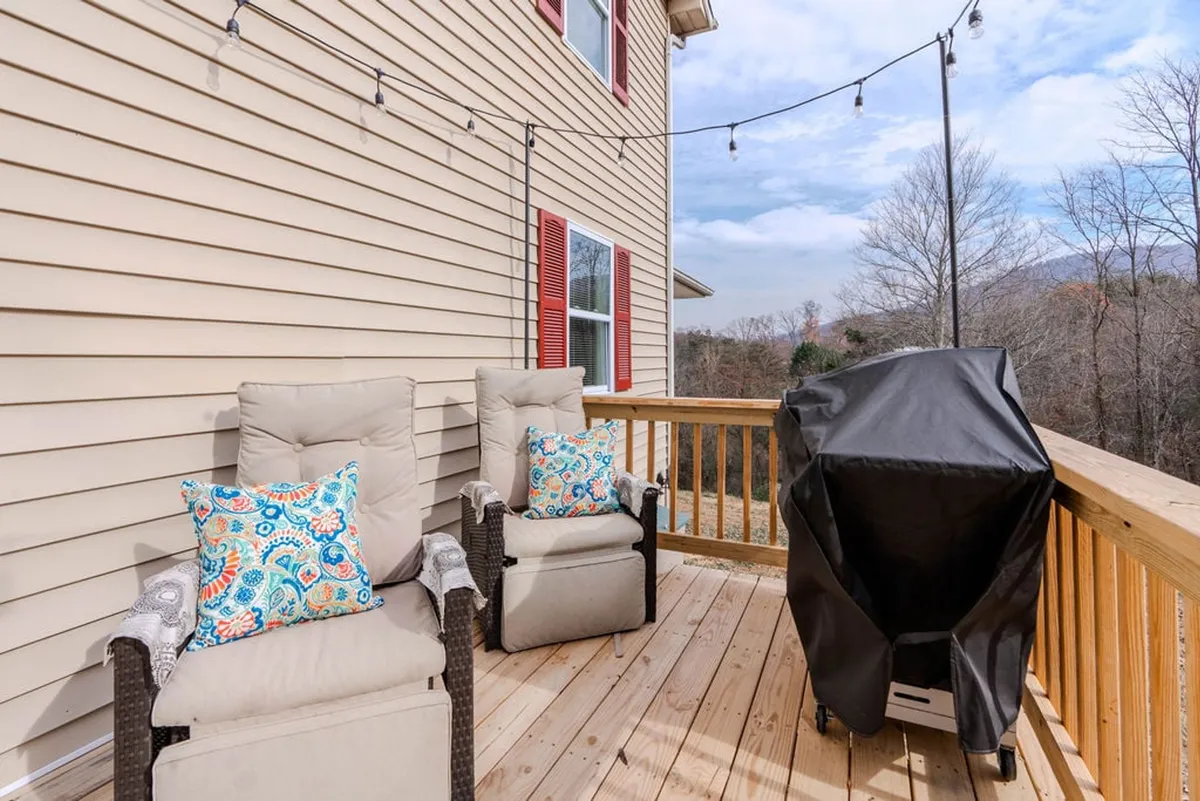 An inviting outdoor patio area featuring comfortable chairs with colorful pillows and a grill, set against a backdrop of trees. The relaxed setup encourages gatherings or quiet moments outdoors, with hints of seasonal beauty visible in the foliage.