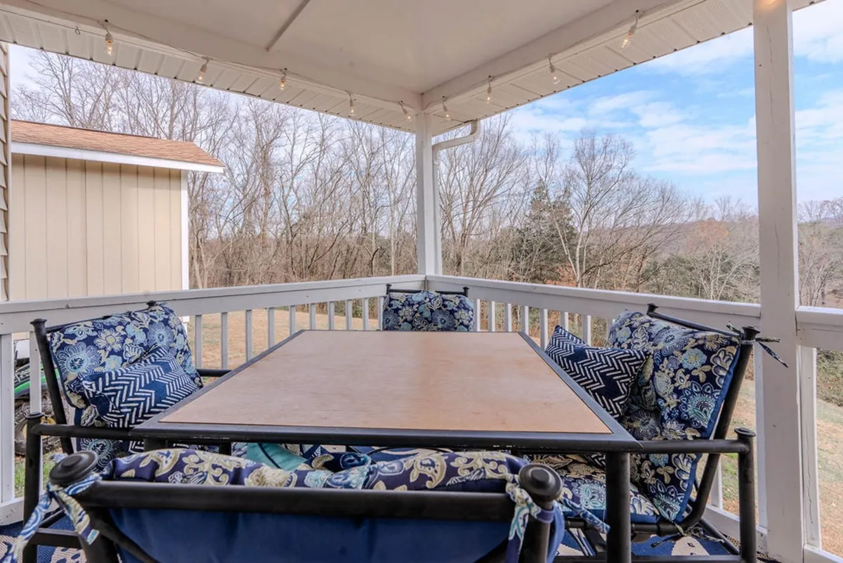 The image captures a cozy outdoor dining area set beneath leafy trees, with a table and decorative cushions inviting group gatherings. The background is lush with nature, evoking relaxation and connection to the outdoors.