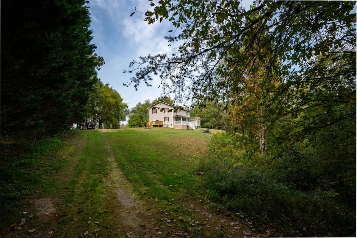 This image presents a broad view of the house’s exterior, with a welcoming porch, surrounding trees, and a visible pathway leading up to the home. The setting feels peaceful and private, ideal for enjoying natural beauty and evening gatherings.