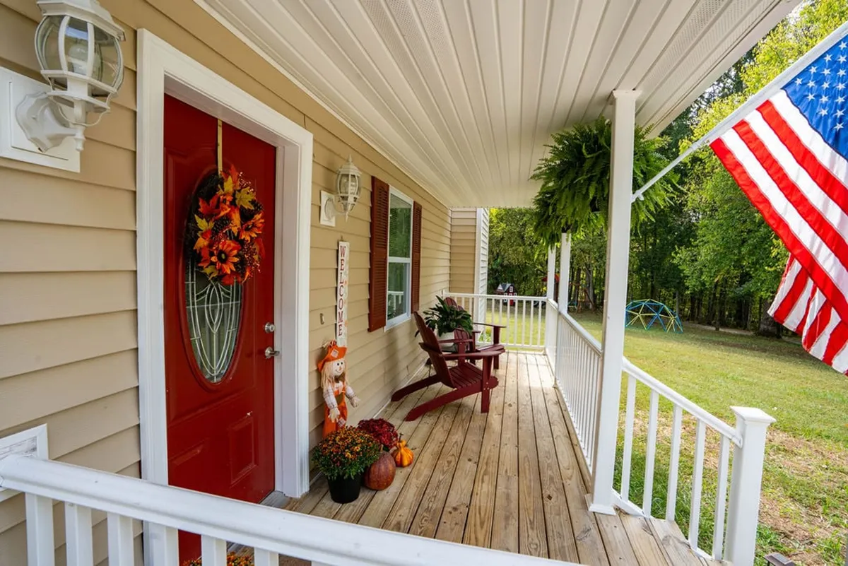 This image captures the exterior of the home, featuring a welcoming red front door adorned with an autumn wreath, flanked by rocking chairs and seasonal fall decorations. The American flag and inviting porch suggest a home that celebrates holidays and welcomes gatherings.