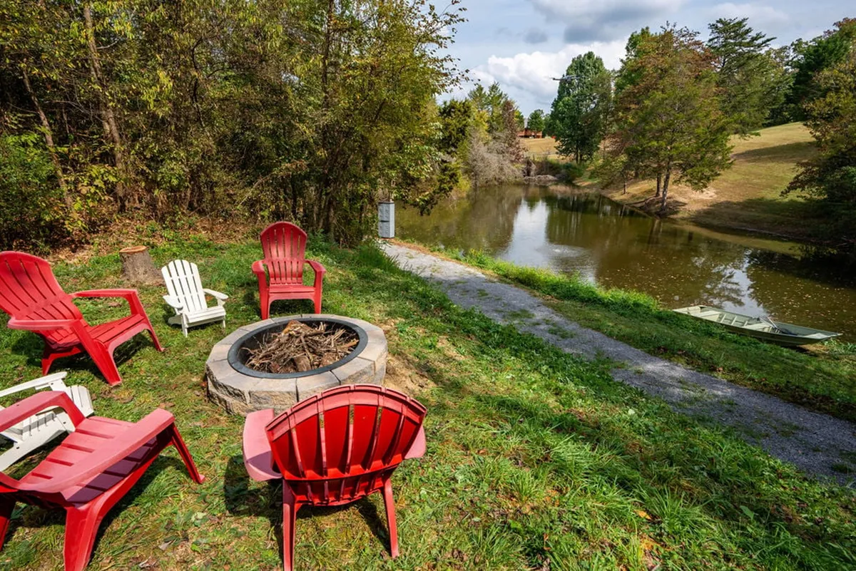 A serene fire pit setup with red chairs arranged around it, positioned beside calm water. The tranquil pond is visible in the background, inviting for boating or fishing. The setting suggests an inviting spot for families and groups to gather under the stars and enjoy the outdoors.